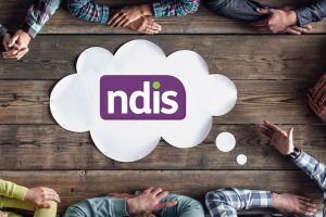 A birds-eye view of the NDIS logo on a paper speech bubble sitting on a wooden desk. Six people sit around the desk. Only their arms are visible.