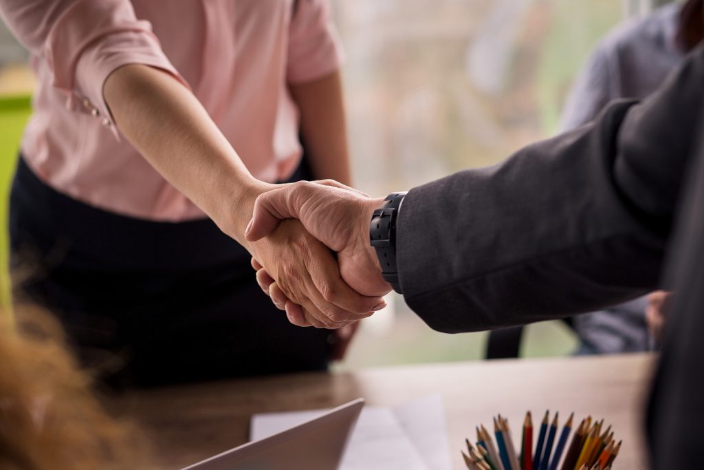 A woman reaches across a desk and shakes hands with another person.