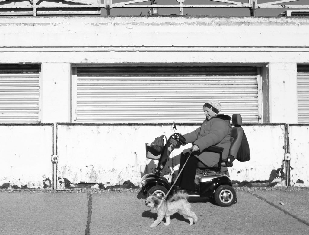  woman wearing a large coat driving past an old building on a mobility scooter. Her dog walks along beside the scooter on a leash.