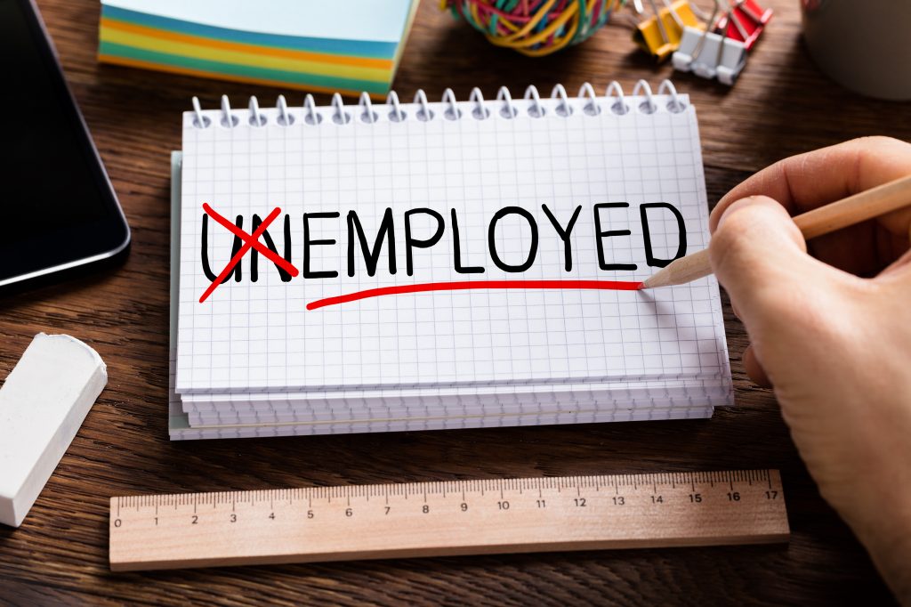 A grid-lined notebook and various office supplies arranged on a desk. The word enemployment is written on the notebook with 'un' crossed out in red. A hand is using a red pen to underline the word 'employed'