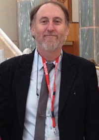 A man with a short grey beard smiles toward the camera. He is wearing a dark suit, light shirt, and a grey tie.