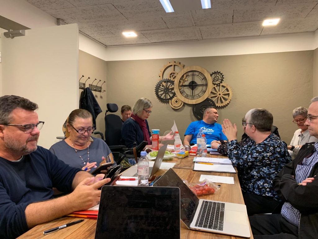 A group of people with disability sitting casually around a table covered in laptops, notebooks and water bottles. They are in a small room with a clockwork sculpture on the wall.