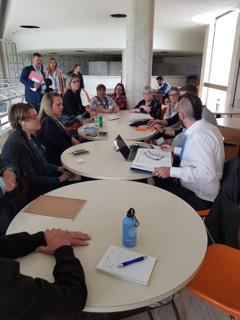 A group of people sitting and standing around a few small round tables pushed together. There are notebooks, laptops and phones on the tables.