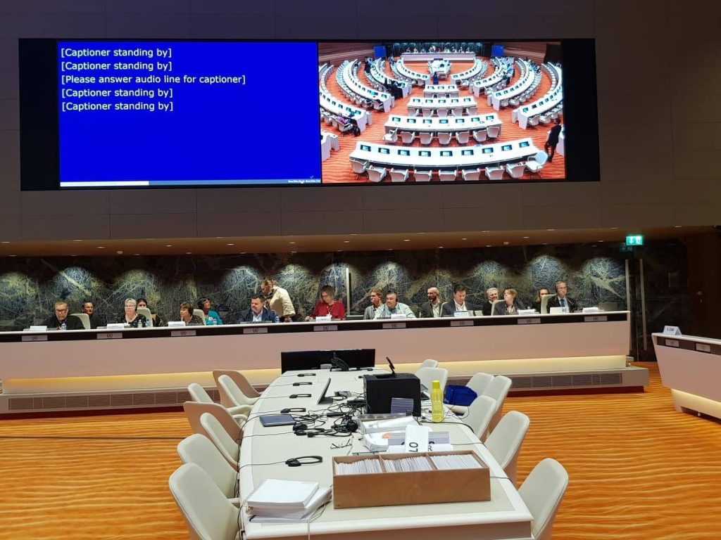 A group of people behind a long panel desk. Behind them is a presentation screen and in front a long table with empty chairs around it and event material on it.