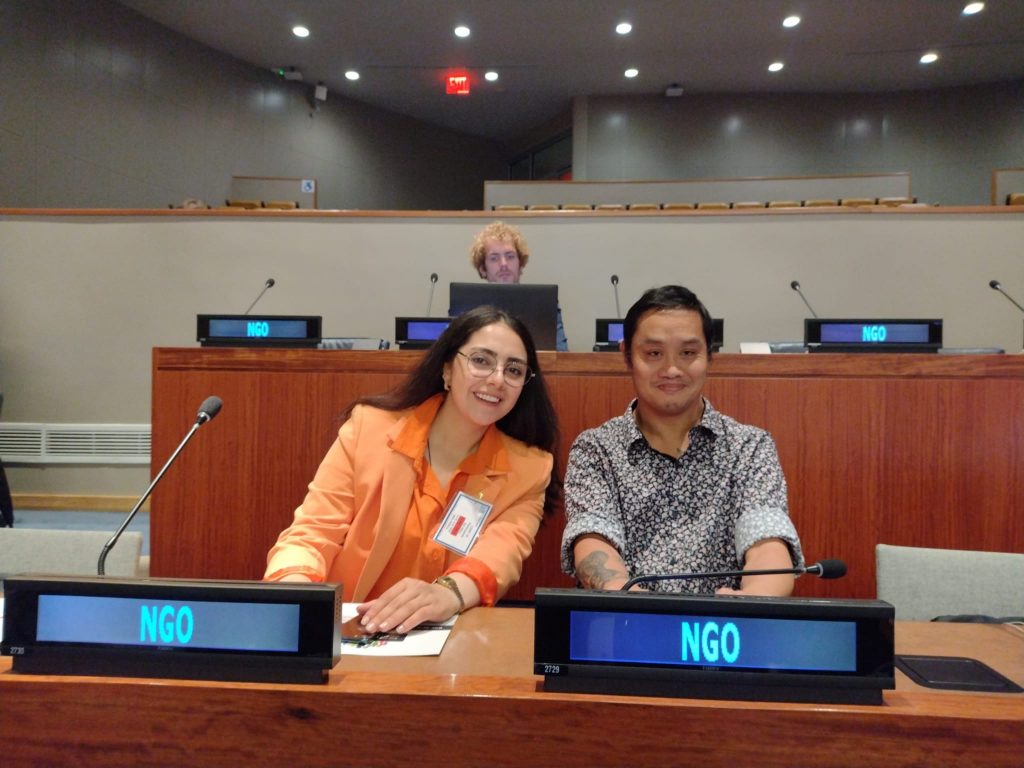 A woman and man smiling at a desk with microphones and signs reading NGO in front of them.
