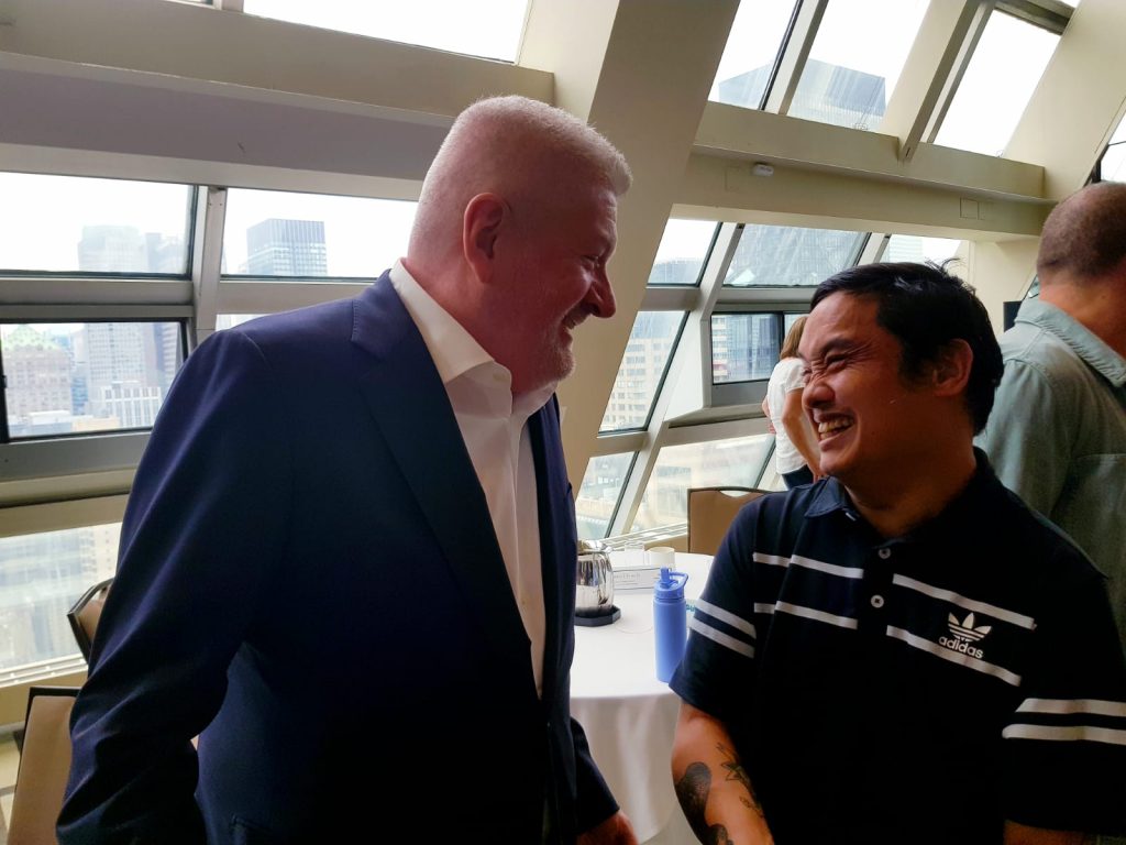 A white-haired white man and an Asian man laughing at the edge of a crowd, in front of a table and a wall of panelled windows.