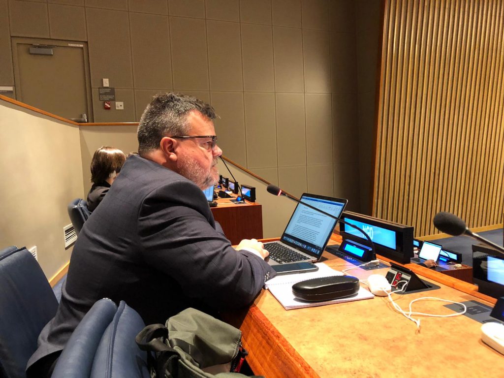 A First Nations man in a suit is speaking into a microphone while seated at a long desk with an open laptop and an NGO sign.