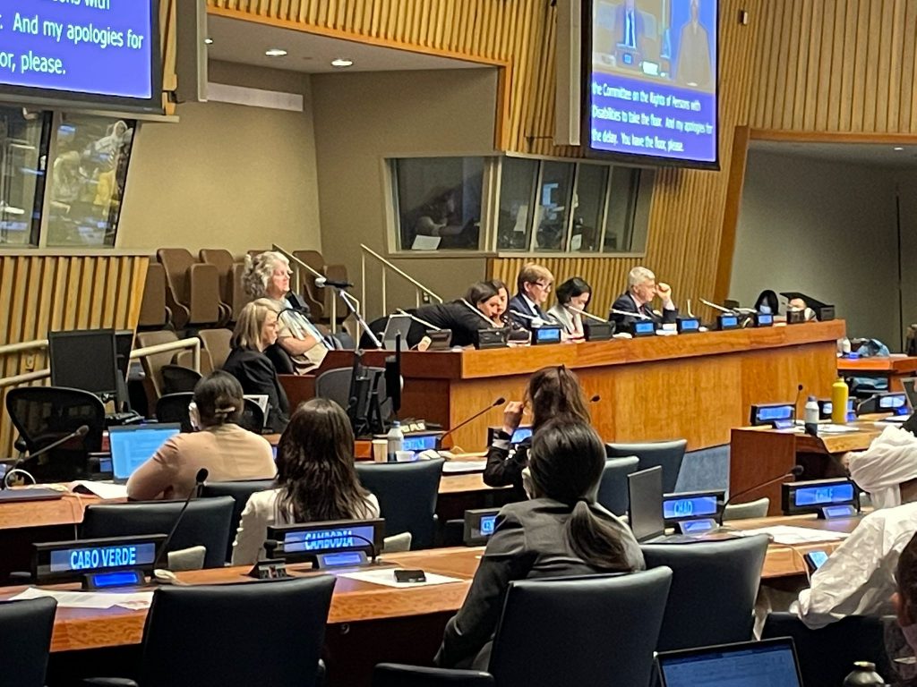 Delegates behind desks with country names on signs listen to a speech by one of the people behind the raised desk at the front of the formal UN meeting room.