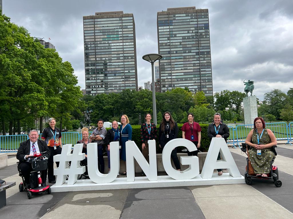 People wearing lanyards pose with a large #UNGA sign in a park with two skyscrapers in the background.