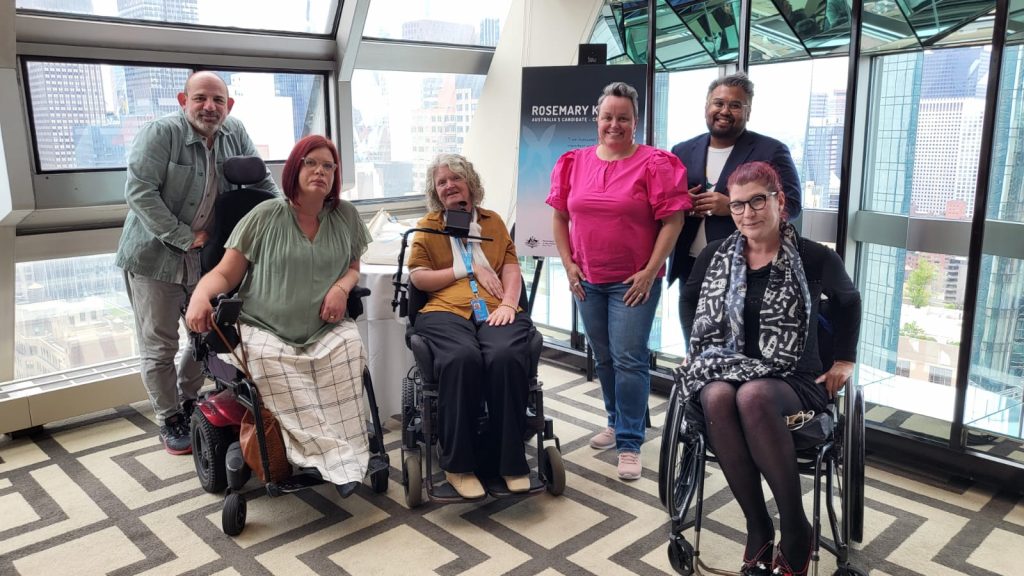 A group of people posing for a photo in a room with large glass walls, high up in a skyscraper. Three of them are in wheelchairs and three are standing. A sign behind them has Rosemary's name on it.