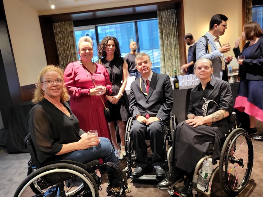 Five well-dressed people posing for a photo indoors, three of them in wheelchairs. Behind them is a large window and a table with bottles and glasses on it.