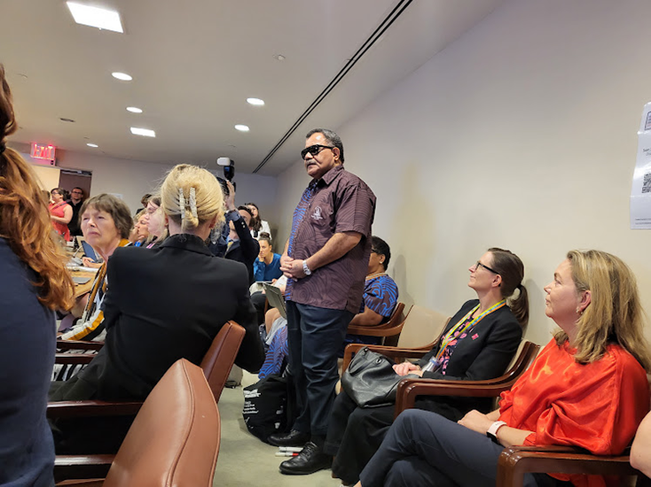 An older man in sunglasses standing up from his chair at the back of an audience in a medium-sized carpeted room.