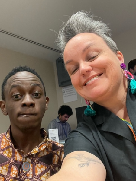 A white woman with short grey hair and  large colourful earrings is taking a selfie with a young black man in a brown and orange patterned shirt. She is smiling and he is raising his eyebrows.