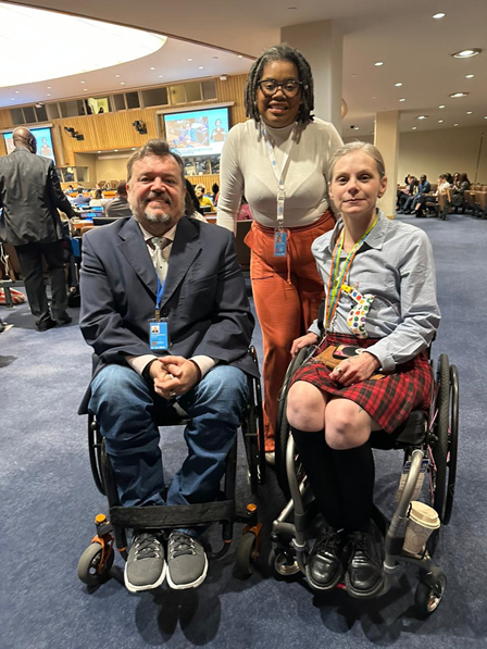 Three people posing at the side of a large open conference chamber. A white man and woman in wheelchairs and a black woman standing behind them. All three are wearing lanyards with nametags.