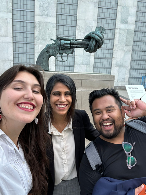 Three people posing for a selfie in front of a large statue of a gun with its barrel twisted into a knot.