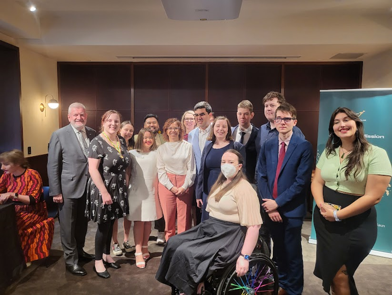 A large group of well-dressed people indoors, mostly young, one in a wheelchair.