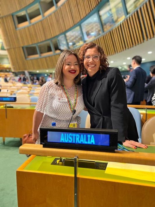 Two women leaning close together and smiling behind the 'Australia' sign on the panel desk. One is wearing a white dress, a rainbow lanyard and bright red lipstick, the other is wearing glasses and a black suit.