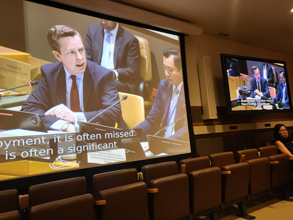 A large projection screen showing a man in a suit speaking while seated at a long desk with microphones.