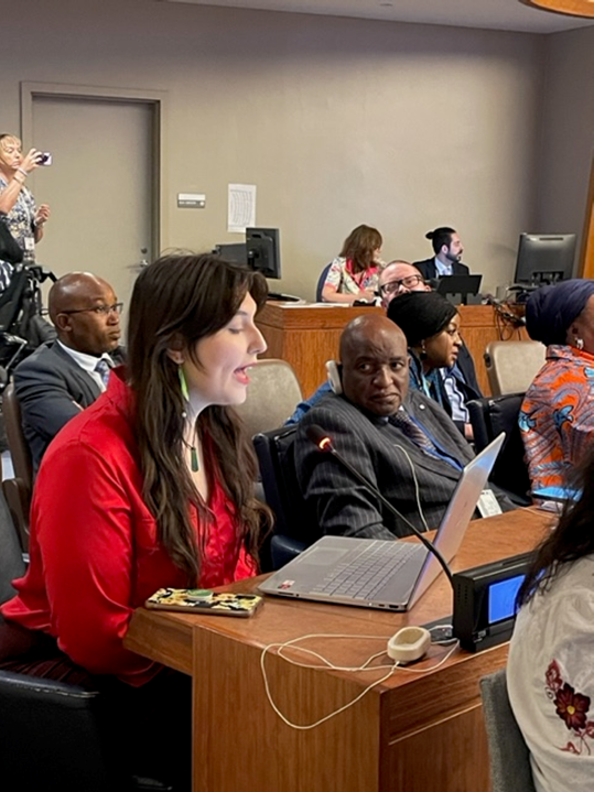 A woman in a bright red blouse looking at a laptop and speaking into a microphone at a desk beside a black man in a suit.