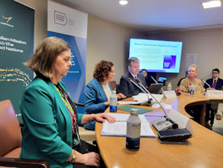 People in suits sitting around a large curved panel with microphones, in a small room with a screen in one corner showing a presentation about Australia's Disability Strategy.