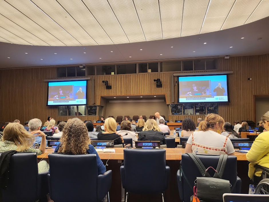 A large room with a panel desk slightly raised at the front and two big screens on the walls showing the current speaker and Auslan interpreter. The audience is seated at long desks with microphones.